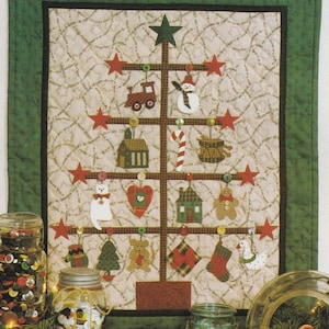 Peut inclure: Une suspension murale en forme de sapin de Noël matelassée avec un cadre en bois brun et une bordure verte. L'arbre est décoré de diverses décorations en tissu, notamment un bonhomme de neige, un bonhomme en pain d'épices, un chat, une maison, une chaussette et une canne en sucre. Les décorations sont attachées à l'arbre avec des boutons.
