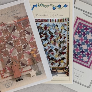 May include: Three quilt pattern booklets are displayed, showcasing various designs. The "Wish and Dream" pattern features a patchwork design with earth-tone colors. "Romantic Charm" displays a colorful patchwork. "Pinwheels In The Attic" has a pink and blue pinwheel design.