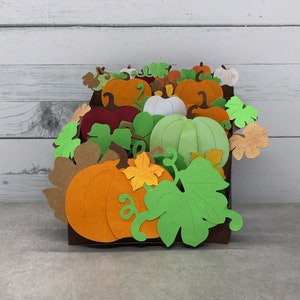May include: A 3D pop-up card featuring a variety of pumpkins in different colors and sizes, including orange, green, white, and red. The pumpkins are surrounded by green leaves and vines, creating a festive autumnal scene.