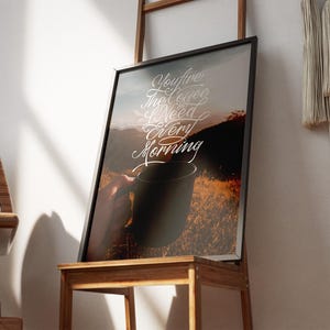 Könnte beinhalten: Gerahmter Kunstdruck mit dem Text "You are the coffee I need every morning". Das Bild zeigt eine Person, die eine Kaffeetasse mit einem Bergpanorama hält, schwarz gerahmt. Auf einem Holzstuhl platziert.