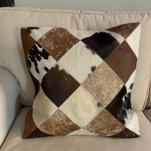 May include: Square decorative pillow with a patchwork design. The pillow features a diamond pattern made of various shades of brown, white, and black cowhide. The pillow is resting on a beige sofa.