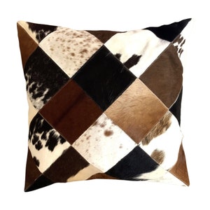 May include: A square throw pillow featuring a patchwork design of cowhide panels. The surface consists of alternating diamond-shaped pieces in various textures and colors, including solid black, dark brown, tan, and white with black or brown spots.