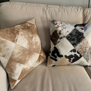 May include: Two square throw pillows featuring a patchwork design of cowhide. The left pillow displays shades of tan and cream, while the right pillow showcases a multi-colored pattern of black, white, brown, and tan patches in a diamond arrangement.