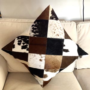 May include: A square throw pillow featuring a patchwork design made of cowhide panels. The surface displays a variety of textures and colors, including solid black, solid brown, white, and spotted patterns in black, brown, and white tones.