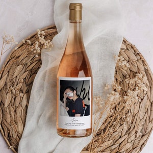 May include: A bottle of ros&eacute; wine with a gold cap and a label featuring a photograph of a person holding a camera. The bottle is set on a woven placemat with a white cloth and dried flowers.