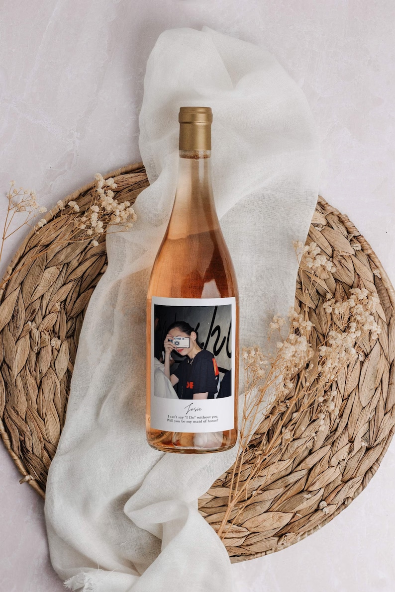 May include: A bottle of ros&eacute; wine with a gold cap and a label featuring a photograph of a person holding a camera. The bottle is set on a woven placemat with a white cloth and dried flowers.