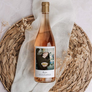May include: A bottle of ros&eacute; wine with a gold cap and a label featuring a photograph of a person. The label includes the name "Megan" and the text "I can't say 'I Do' without you. Will you be my maid of honor?" The bottle rests on a woven placemat.