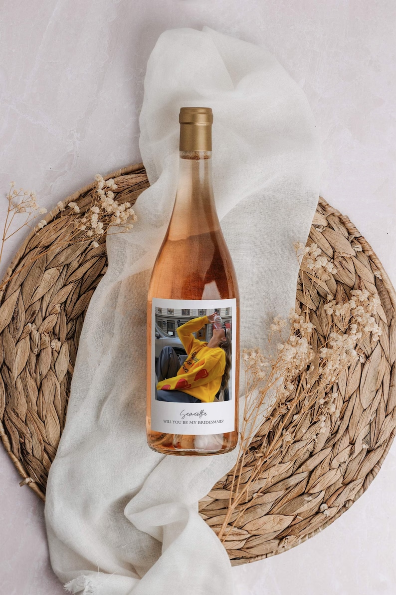 Op de afbeelding: Een fles ros&eacute; wijn met een gepersonaliseerd etiket. Het etiket bevat een foto van een persoon die een geel sweatshirt draagt met rode harten en de tekst "Wil je mijn bruidsmeisje zijn?" De tekst "Savannah" is in cursief geschreven onderaan het etiket.
