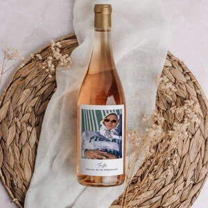 Op de afbeelding: Een fles ros&eacute; wijn met een gepersonaliseerd etiket met een foto van een persoon met een zonnebril en een groen-wit gestreept shirt. Het etiket luidt "Wil je mijn bruidsmeisje zijn?" met de naam "Trista" eronder.