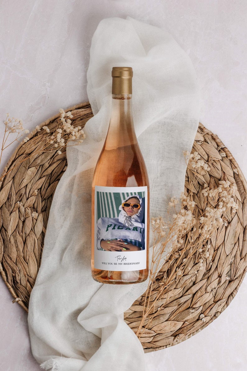Op de afbeelding: Een fles ros&eacute; wijn met een gepersonaliseerd etiket met een foto van een persoon met een zonnebril en een groen-wit gestreept shirt. Het etiket luidt "Wil je mijn bruidsmeisje zijn?" met de naam "Trista" eronder.