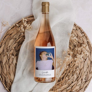 May include: A bottle of ros&eacute; wine with a gold cap and a label featuring a photo of a person drinking from a can. The label includes the name "Scarlette" and the text "I can't say 'I Do' without you. Will you be my maid of honor?" The bottle rests on a woven placemat.