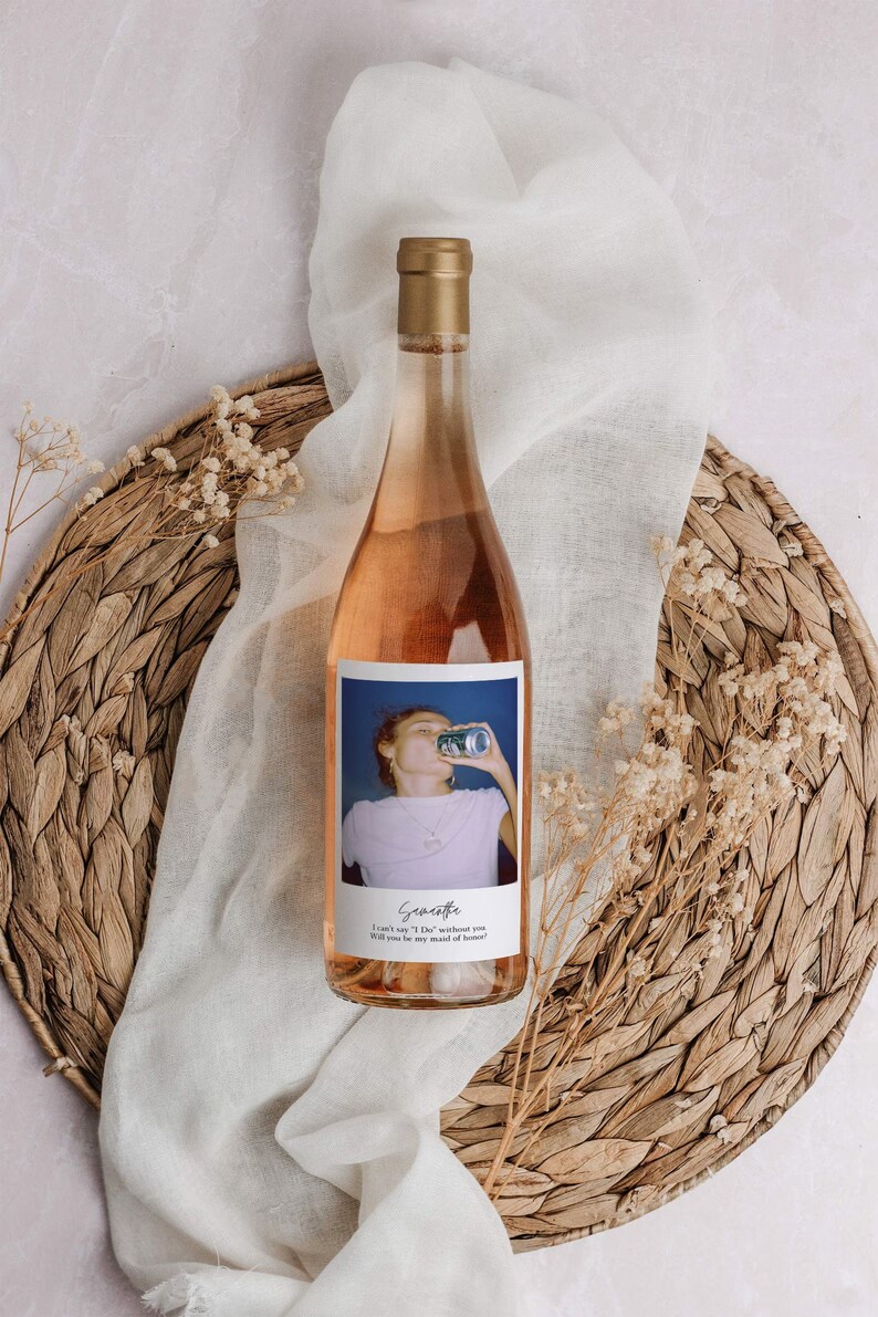 May include: A bottle of ros&eacute; wine with a gold cap and a label featuring a photo of a person drinking from a can. The label includes the name "Scarlette" and the text "I can't say 'I Do' without you. Will you be my maid of honor?" The bottle rests on a woven placemat.