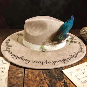 May include: A tan fedora hat with a white band, adorned with a blue and green feather and a small decorative charm. The hat features the text "Moderns and laughter, love, blend of perfect sisters" around the brim. The hat is on a wooden surface.