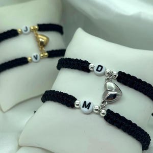 May include: Two matching black braided bracelets with magnetic heart charms. One bracelet has the letters "J" and "V" on white beads, the other has "D" and "M". The bracelets are displayed on white pillows.