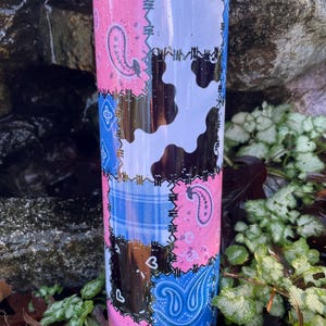 May include: A stainless steel tumbler with a patchwork design. The tumbler features pink, blue, and black paisley, plaid, and cow print patterns. The design is reminiscent of a quilt. The tumbler is cylindrical and appears to be insulated.