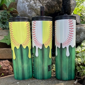 May include: Three insulated tumblers with black lids. The tumblers feature a baseball design with red stitching on a white background, yellow dripping paint, and green vertical stripes. One tumbler has the letter "N".