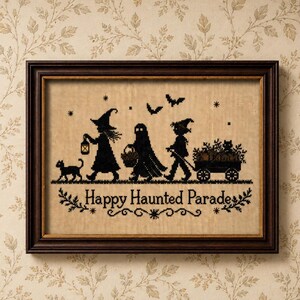 May include: A framed cross-stitch embroidery on tan fabric featuring a black silhouette design of a witch, a ghost, and a child pulling a wagon filled with pumpkins and a cat. The text "Happy Haunted Parade" is stitched in black below the figures.