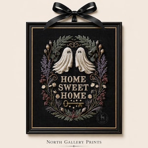 May include: A framed embroidery piece on black fabric featuring two white ghosts, a floral wreath, and the text "HOME SWEET HOME" with a key icon. The rectangular black frame is topped with a black satin ribbon bow. The design includes botanical elements.