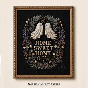 May include: A framed embroidery art piece on black fabric featuring two white ghosts, a central gold key, and the text "HOME SWEET HOME" surrounded by a wreath of dried flowers, seed pods, and greenery in muted tones of purple, green, and brown.
