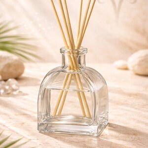 Puede incluir: Un frasco difusor de aromaterapia de vidrio transparente con varillas de madera. El frasco tiene una base cuadrada y una parte superior redondeada. Las varillas son de color marrón claro. El fondo es de color beige claro con sombras y un símbolo del zodiaco.