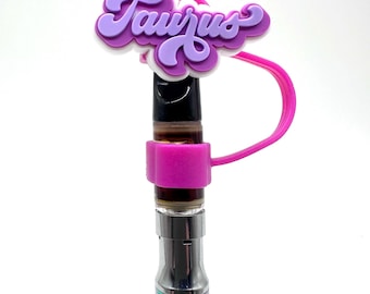 May include: A vape pen with a purple and white Taurus charm. The pen has a metallic, iridescent finish. The charm is attached to a pink silicone sleeve that wraps around the pen's cartridge.