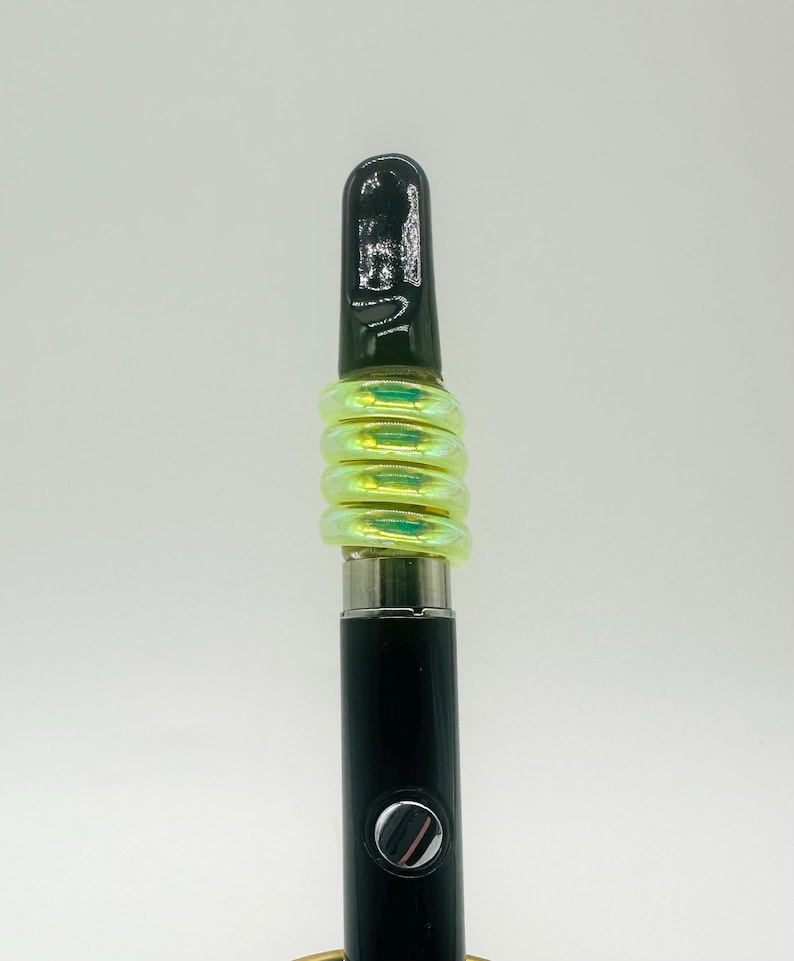 May include: A black vape pen with a green iridescent silicone sleeve. The sleeve is made up of multiple rings.