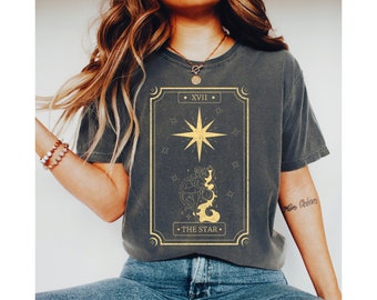 May include: Dark gray t-shirt featuring a gold-colored tarot card design. The design includes a star, the Roman numeral XVII, and the words "THE STAR". The shirt is a relaxed fit and has short sleeves.