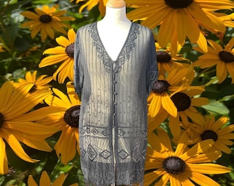 May include: A sheer, charcoal gray duster cardigan featuring intricate beadwork in geometric patterns, a deep V-neckline, short sleeves, and a front button closure with a fringed hemline, displayed on a mannequin against a backdrop of yellow flowers.