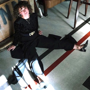 May include: A woman wearing a black velvet top with a gold belt and black pants sits on a floor with red and black lines. She is wearing gold hoop earrings and black shoes.