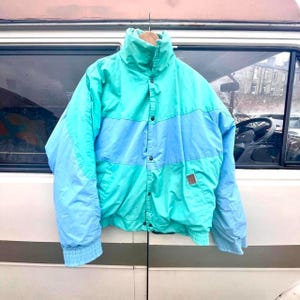 May include: A color-blocked jacket featuring a high collar, long sleeves with ribbed cuffs, and a front button closure. The design alternates between panels of bright turquoise and light blue, with a small brown rectangular patch on the lower left.