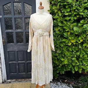 May include: A cream-coloured vintage dress with long sleeves and a floral design. The dress has a V-neck, a belted waist with a circular buckle, and a flowing skirt. The dress is displayed on a mannequin, with a dark door in the background.
