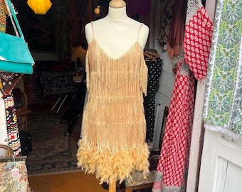 May include: A sleeveless gold dress featuring multiple horizontal layers of fringe covering the entire bodice and skirt, finished with a dense trim of matching gold feathers along the hemline, displayed on a mannequin with thin shoulder straps.