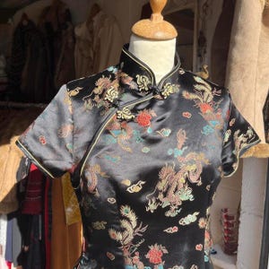 May include: A black short-sleeved blouse featuring a mandarin collar and a diagonal front closure with fabric toggles. The garment is decorated with a colorful embroidered pattern of dragons, phoenixes, and floral motifs in red, gold, and blue.