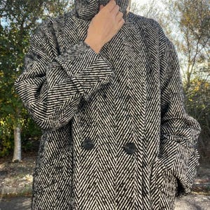 May include: A double-breasted coat featuring a black and white herringbone pattern. The coat has a high collar, long sleeves with cuffs, and two large black buttons centered on the front. The fabric has a textured appearance throughout.