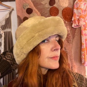 May include: A beige faux fur bucket hat with a thick, rounded crown and a wide, soft brim. The hat features a plush, uniform texture throughout, providing a full, rounded silhouette that sits comfortably on the head with a dense, fuzzy finish.