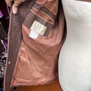 May include: A brown leather jacket with a smooth brown interior lining. The interior features a rectangular label with the text Real Leather and a smaller white care tag below it. The jacket has a single visible gold-toned metal button on the front.