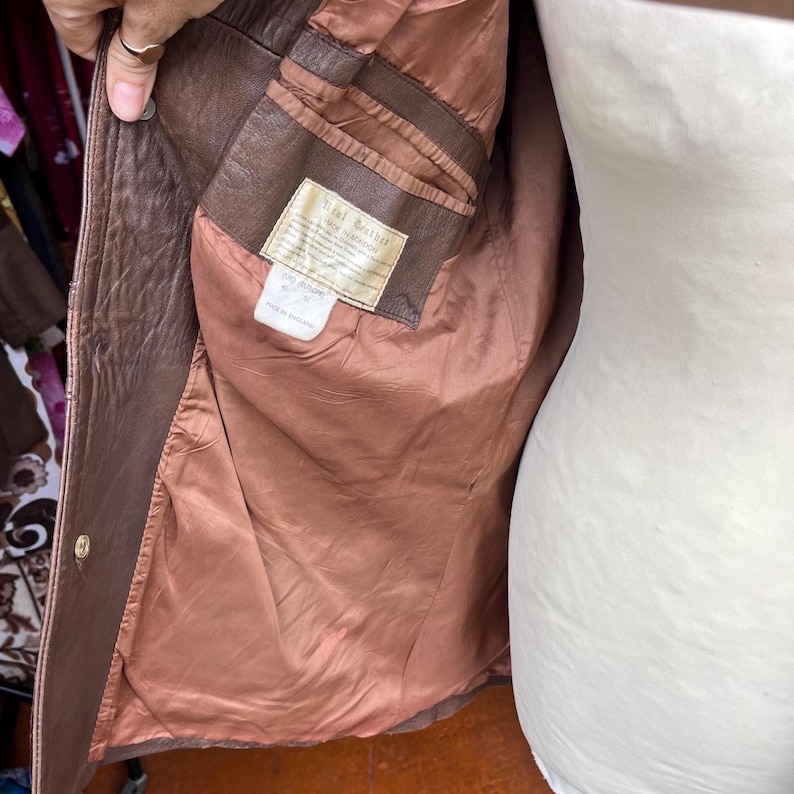 May include: A brown leather jacket with a smooth brown interior lining. The interior features a rectangular label with the text Real Leather and a smaller white care tag below it. The jacket has a single visible gold-toned metal button on the front.