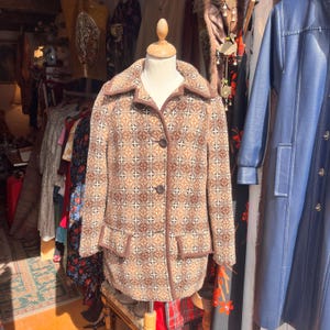 May include: A long-sleeved jacket featuring a brown, tan, and cream geometric pattern. The jacket has a wide collar, two front buttons, and two flap pockets at the waist. The edges are finished with solid brown trim, and the fabric has a textured appearance.