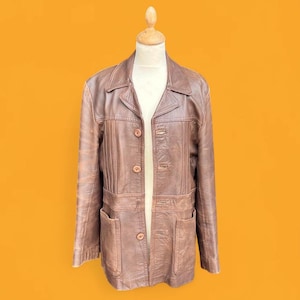 May include: A brown leather jacket featuring a collared neckline, long sleeves, and a button-front closure. The garment includes vertical stitched paneling on the chest and two large patch pockets at the waist, displayed on a mannequin form.