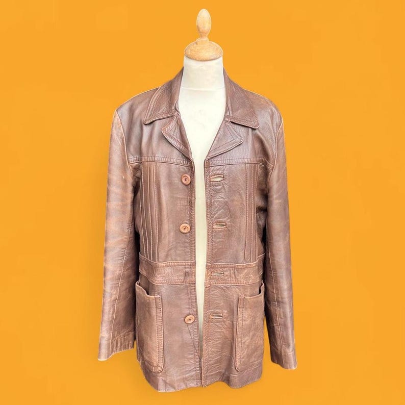 May include: A brown leather jacket featuring a collared neckline, long sleeves, and a button-front closure. The garment includes vertical stitched paneling on the chest and two large patch pockets at the waist, displayed on a mannequin form.