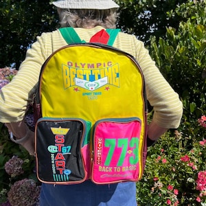 May include: A retro yellow backpack with multiple pockets and vibrant details. The backpack displays the text "OLYMPIC BEST TIME" and "SPORT TIMES 1958". Additional pockets feature the text "START" and "773 BACK TO BASIC".