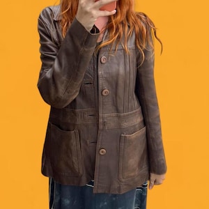 May include: A brown leather jacket featuring a collared neckline, long sleeves, and a front button closure. The garment includes vertical stitched detailing on the upper chest and two large patch pockets on the lower front section of the jacket.