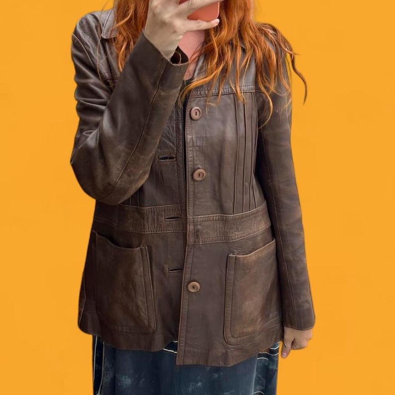 May include: A brown leather jacket featuring a collared neckline, long sleeves, and a front button closure. The garment includes vertical stitched detailing on the upper chest and two large patch pockets on the lower front section of the jacket.