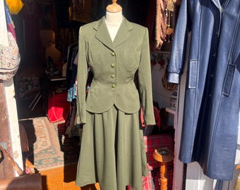 May include: An olive green two-piece suit consisting of a long-sleeved blazer with a notched lapel, four front buttons, and two pockets, paired with a matching midi-length skirt that features a flared silhouette and a smooth, solid-colored fabric.