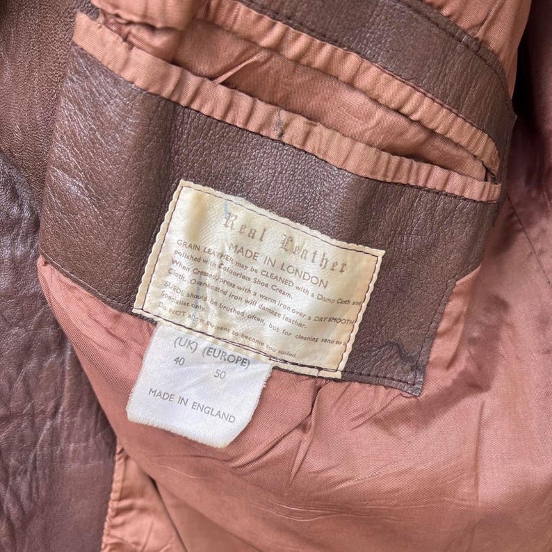 May include: A brown leather jacket interior featuring a cream-colored label with the text Real Leather, Made in London, and care instructions. A secondary white tag displays the sizes UK 40 and Europe 50, with the text Made in England printed below.