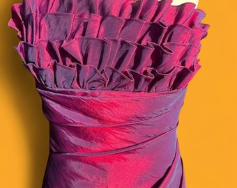 May include: A strapless dress in a shimmering iridescent purple and magenta fabric. The bodice features multiple horizontal layers of ruffled fabric, while the lower section displays diagonal ruching across the front of the garment.