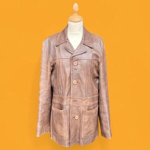 May include: A brown leather jacket featuring a pointed collar, long sleeves, and a front button closure with four visible buttons. The jacket includes vertical seam detailing across the chest and two large patch pockets on the lower front section.