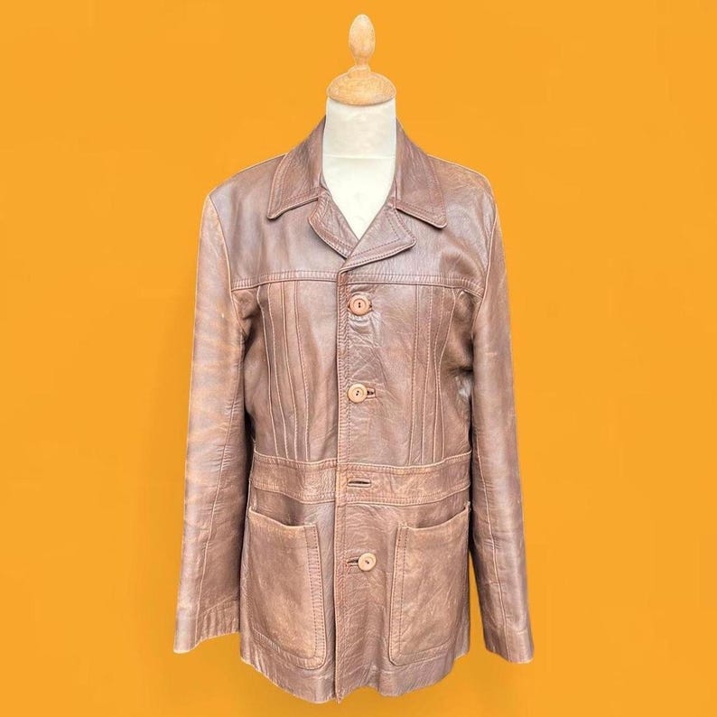 May include: A brown leather jacket featuring a pointed collar, long sleeves, and a front button closure with four visible buttons. The jacket includes vertical seam detailing across the chest and two large patch pockets on the lower front section.