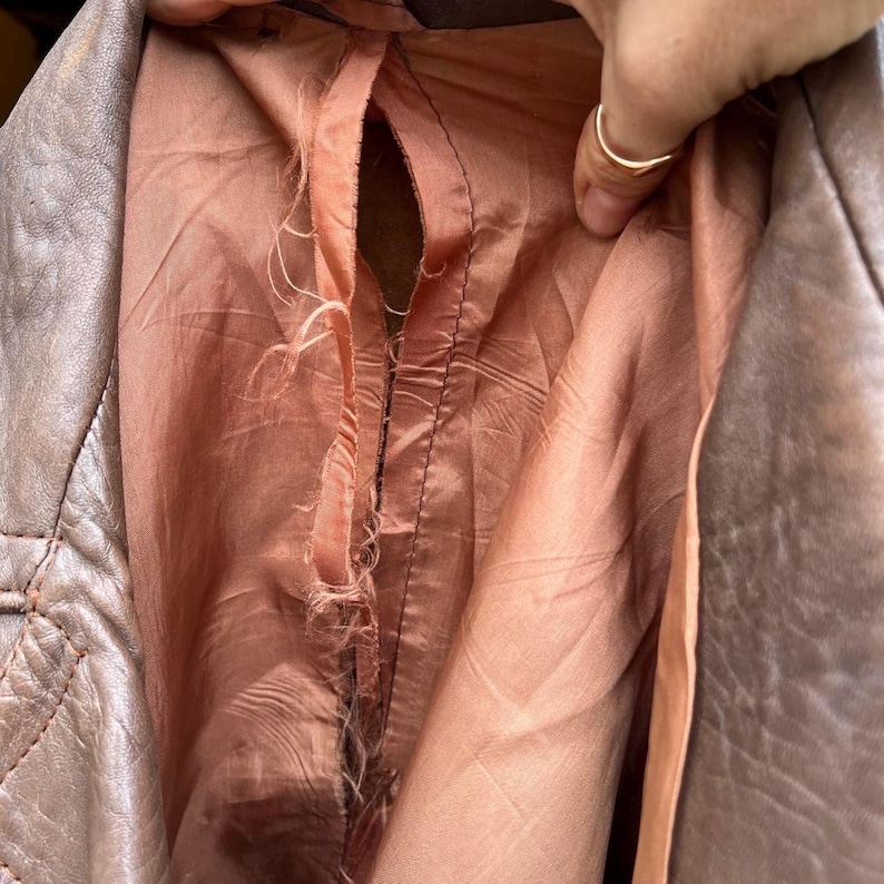 May include: A brown leather jacket with a visible tear along the interior seam. The lining is a dusty rose color and shows significant fraying and loose threads where the fabric has separated from the stitching along the central vertical seam.