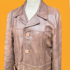 May include: A brown leather jacket featuring a pointed collar, long sleeves, and a front button closure with two visible round buttons. The garment displays vertical stitched paneling details across the chest and a textured finish throughout.